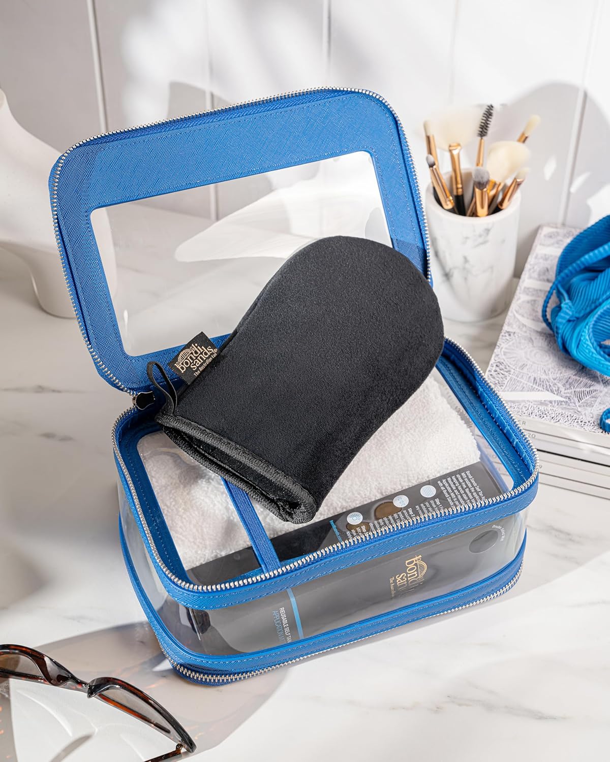Bondi Sands tanning mitt packed in a clear blue-trimmed travel case with a white towel on a marble bathroom counter