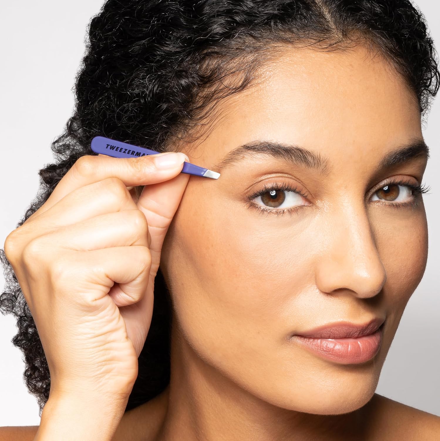 A woman using the purple Tweezerman Mini Slant tweezer to shape her eyebrow