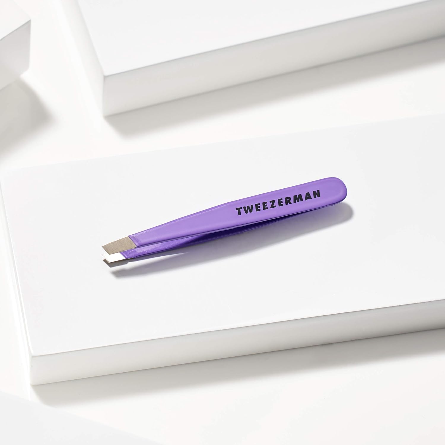 The periwinkle Tweezerman Mini Slant tweezer resting on a white block with the branding visible