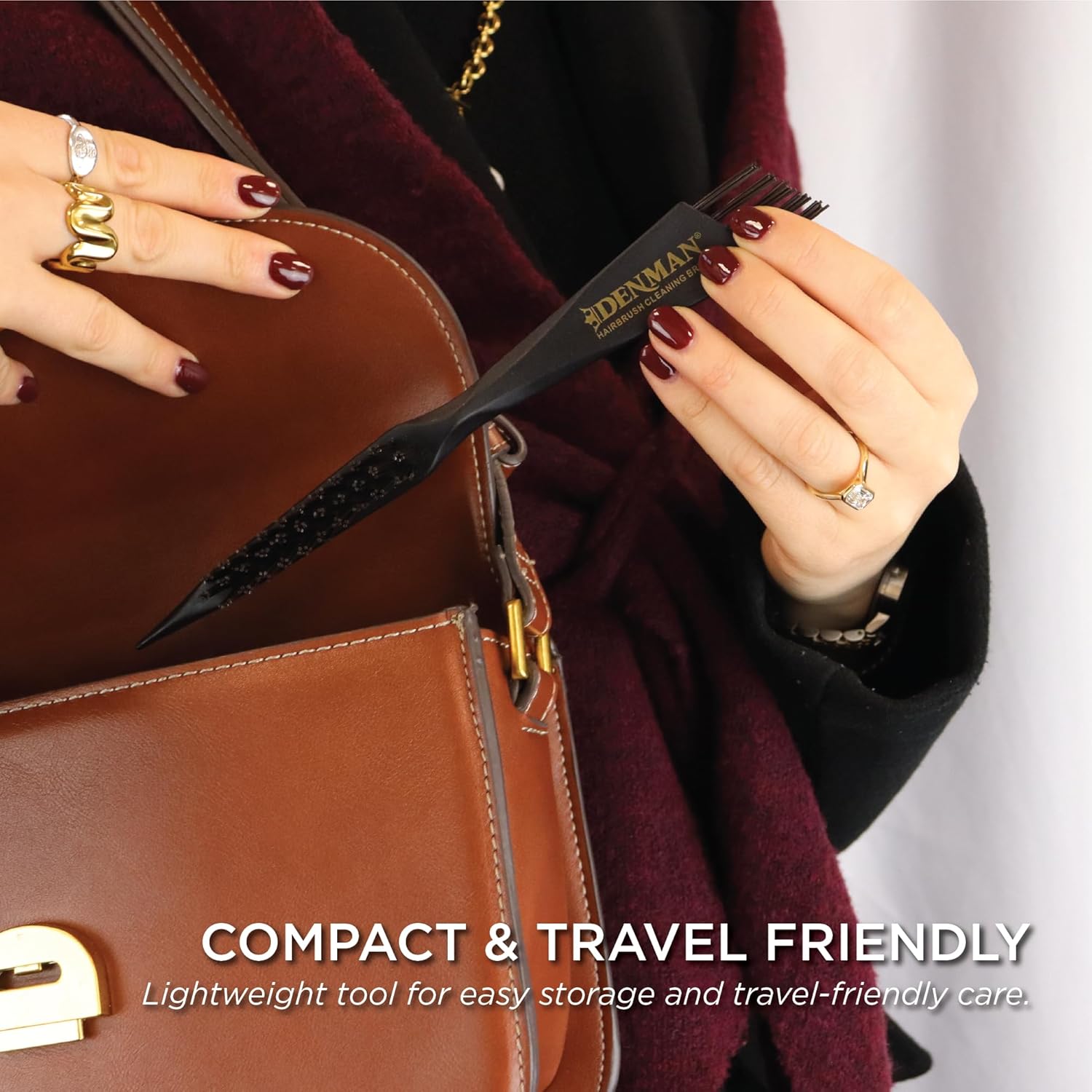 The compact Denman DCB1 cleaning brush held beside a brown leather handbag, showing its travel-friendly size