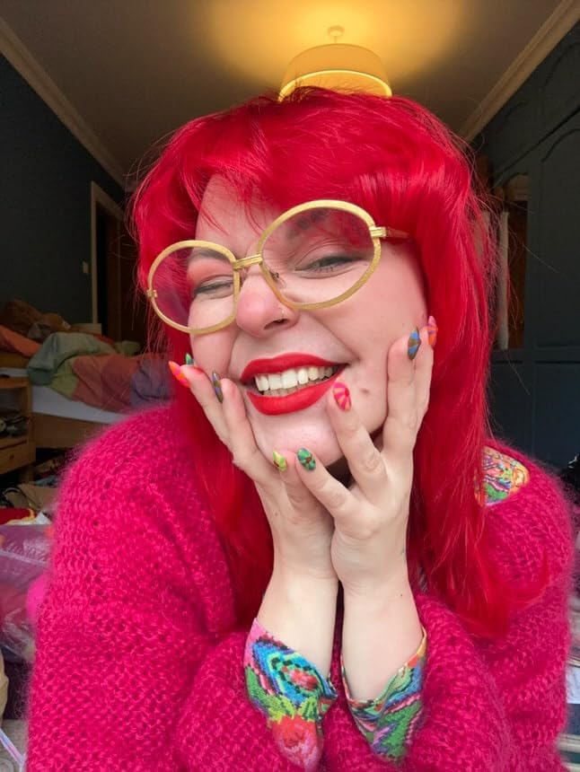 Smiling woman with bright vermillion red dyed hair wearing round gold glasses and a pink jumper