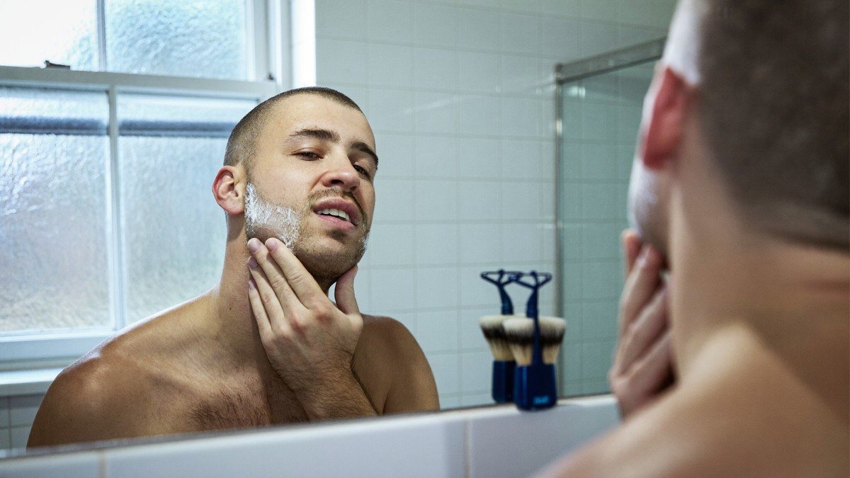 A man at a bathroom mirror on a cold morning with shaving foam along his jaw, checking his face in the daylight coming through a frosted window