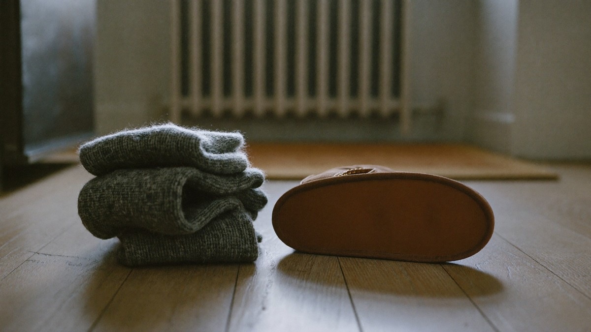 A folded stack of thick grey wool socks and a leather winter boot on a wooden hallway floor beside a radiator on an autumn morning
