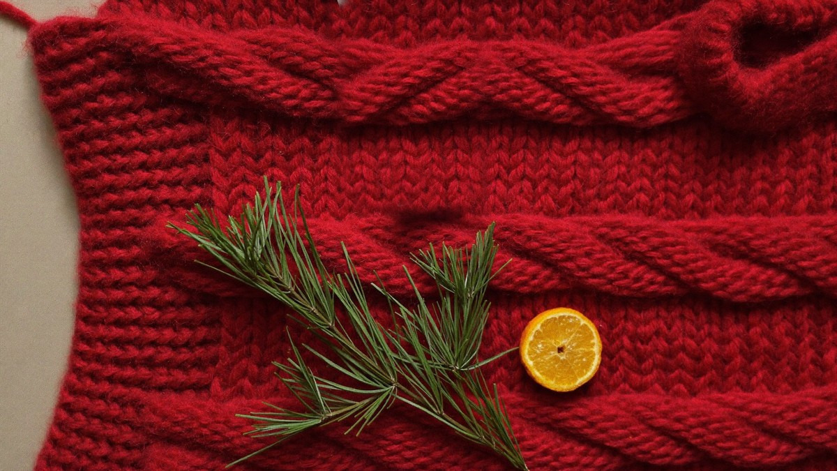 Close-up of a chunky deep red cable-knit wool Christmas jumper with a sprig of pine and a dried orange slice resting on it, ready for the December jumper season