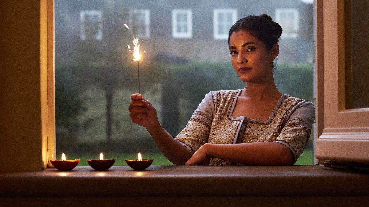 A woman in an embroidered festive kurta holding a lit sparkler at a UK doorway, with three clay diyas burning on the sill on a hazy Diwali evening in November