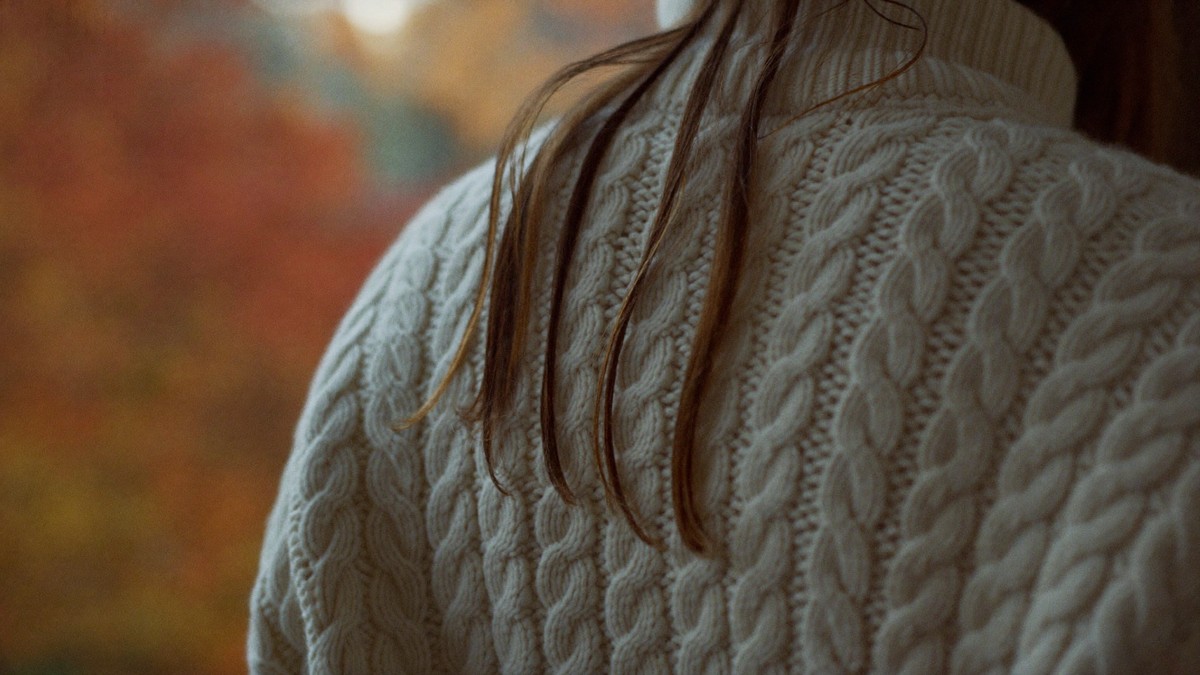 Long brown hair draped over the shoulder of a cream cable-knit roll-neck jumper, with autumn leaves blurred in the background